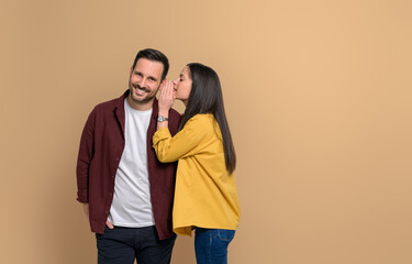 Wall Mural - Side view of young woman covering ear of handsome smiling boyfriend while telling him gossip on isolated beige background. Couple standing together and sharing secrets with each other