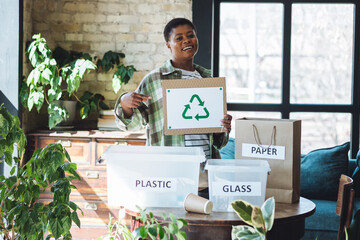 Young plus size African American woman doing waste sorting at home, care about planet, ecology. Containers for recyclable waste: plastic, glass, paper. Holding symbol of recycling, environment protect