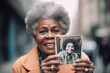 © Trinette Reed - Senior black woman holding up a vintage photo of herself when she was younger age outdoors. Generative AI.