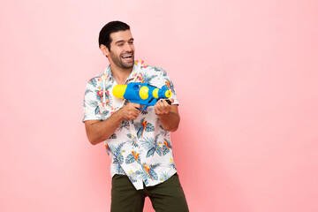 Handsome smiling young Caucasian tourist man playing with water gun in studio pink background for Songkran festival concept in Thailand and southeast Asia