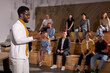 © Roman - Side View Of Nice African American Teacher In White Clothing Lecturing Presenting Speech Gesturing With Hands, Discussing Important Theme With Students, Group Of People In The Background