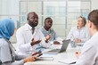 © AnnaStills - African American doctor in white coat sitting at table with laptop and discussing the way of treatment with other colleagues at meeting