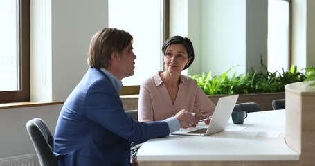 Wall Mural - Mature couple of coworkers talking at workplace with laptop. Businesswoman negotiating with deal partner, colleague, employee, sitting at table, telling work tasks, speaking