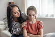Mother comforts upset daughter that sits with arms crossed on a couch.