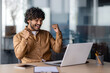 Successful hispanic businessman programmer celebrating victory and triumph, man inside office sitting at table and working using laptop, holding hands up gesture of achieving good results.