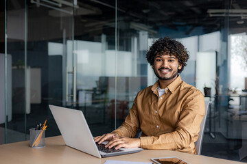 Portrait of young successful Indian man at workplace inside office, businessman smiling and looking at camera, man at work using laptop, programmer with curly hair coding software.