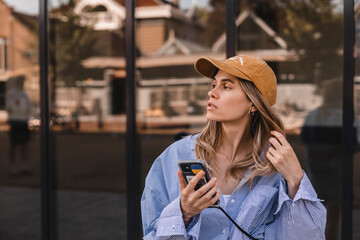 Wall Mural - Close up portrait of a cheerful young woman hold mobile phone outdoors, fix her hair in the city walking near office. Happy woman wear stripped shirt and beige hat cap. Female look at side.