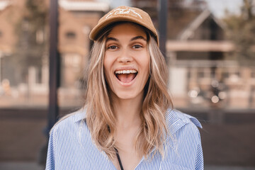 Canvas Print - Cute young woman with a lovely sense of humor standing leaning against a glass window exterior with copy space in an urban street laughing at the camera. Attractive blonde girl wear shirt and cap.