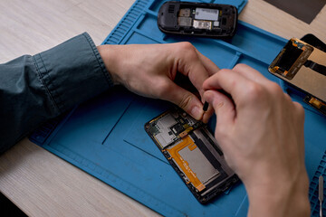 Wall Mural - close-up of the cropped repairman's hands unscrewing screws from smartphone case with screwdriver for repair and maintenance. repair and maintenance of mobile phones. selective focus. top view