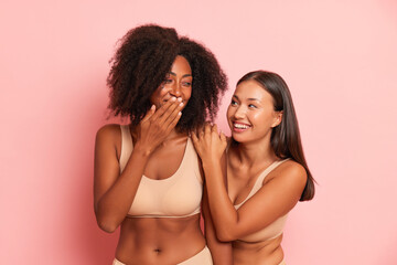 Two smiling young women in beige sport bra are standing against studio background, black skinned girl covers her mouth with hand and smiles, fun time concept, copy space, high quality photo