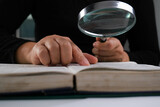Fototapeta Uliczki - Close-up of a woman looking through a magnifying glass at a textbook. Magnifying glass in hand and open book on table. Education and research concept.