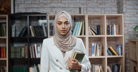 Wall Mural - Portrait of female muslim student, wearing hijab, who is at campus library and confidently looking at camera - modern Islam concept