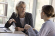 © fizkes - Two young and mature business colleagues women talking at work table, discussing project strategy, plan. Mature professional speaking to coworker, training intern, new employee