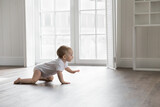 Fototapeta  - Adorable baby wearing white bodysuit, crawling on knees on floor at home. Curious active little infant child learning to move on warm heating safe floor, passing by window in background