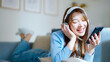 © Monster Ztudio - Happy young asian woman relaxing at home. Female smile sitting on sofa and holding mobile smartphone. Girl using video call to friend. Listening to music