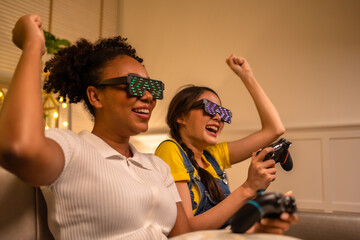 Wall Mural - Young  asian and african woman playing video games at home living room at night.Lesbian couples or lgbt activities on vacation inside the room.