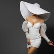 Fashion Woman in Big White Hat hiding Face. Elegant Bride in Mini Dress. Beautiful Lady in Wedding Gown. Stylish Glamour Girl in Sexy Party Dress over Gray Background