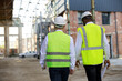 © sakoqwakestock - Back view of builder and architect inspecting building area while walking at construction site. Multiethnic partners wearing hardhats and reflective vest for protecting from accidents.