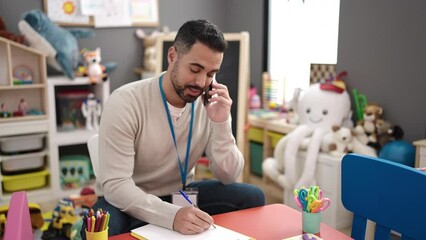 Canvas Print - Young hispanic man preschool teacher writing on document talking on smartphone at kindergarten