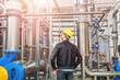 © Olha - A young engineer in a yellow hard hat and jacket at the factory checks the operation of pipelines that are part of an industrial plant for the treatment of contaminated water.