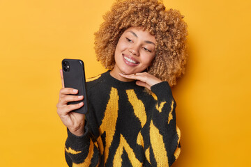 Wall Mural - studio shot of smiling glad european woman with curly hair touches chin gently takes selfie photo vi