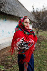 Wall Mural - Charming woman in traditional ukrainian handkerchief, necklace and embroidered dress standing at background of decorated hut with thatched roof. Ukraine, style, folk, ethnic culture