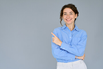 Happy young smiling professional business woman wearing blue shirt looking at camera pointing finger away at copy space showing aside presenting advertising offer standing isolated at gray background.