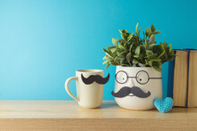 Happy Father's Day Concept With Cute Funny Plant, Coffee Cup And Heart Shape On Wooden Table Over Blue Background