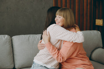 Two lovely happy adult women mature mom young kid wear casual clothes close eyes hug embrace sit on gray sofa couch stay at home flat rest relax spend free spare time in living room. Family concept.