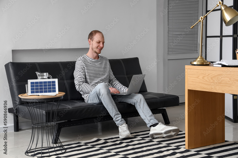 Young man using laptop and portable solar panel charging mobile phone on table at home