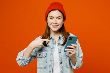 Young smiling fun woman wear denim shirt white t-shirt red hat hold in hand use point index finger on mobile cell phone browsing surfing internet isolated on plain orange background studio portrait.