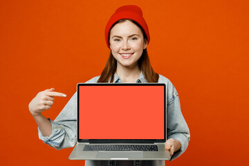 Young fun smiling happy IT woman wears denim shirt white t-shirt red hat hold use work point finger on laptop pc computer with blank screen workspace area isolated on plain orange background studio.