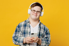 Young Smiling Man With Down Syndrome Wear Glasses Casual Clothes Headphones Listen To Music Use Mobile Cell Phone Isolated On Pastel Plain Yellow Color Background. Genetic Disease World Day Concept.