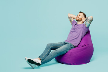 Wall Mural - Full body side view happy young man he wears purple t-shirt sit in bag chair hold hands behind neck isolated on plain pastel light blue cyan background studio portrait Tattoo translates life is fight