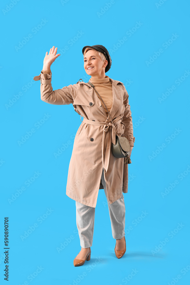 Stylish mature woman waving hand on light blue background