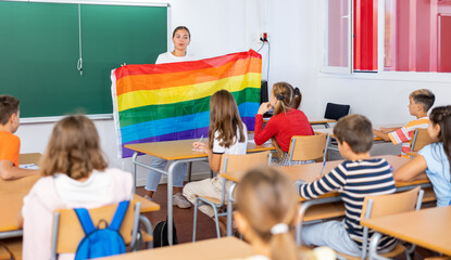 Wall Mural - Young woman teacher holding rainbow flag while explaining lesson about LGBTQ people to young girls and boys.