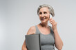 © LIGHTFIELD STUDIOS - overjoyed grandma holding fitness mat and talking on mobile phone isolated on grey.