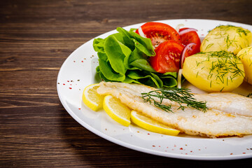 Fish dish - fried cod with boiled potatoes and fresh vegetables on wooden table
