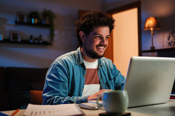 Happy freelancer man working at home with a laptop on a desk at late night. Young guy smiling and browsing on internet using a computer at living room. Entrepreneur looking bank finances and invests