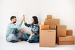 Laughing young couple giving high five sitting on floor in their new home with moving boxes on white background