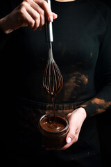 Wall Mural - A jar of melted chocolate in the hands of a chocolatier. Kitchen whisk. On a black background.