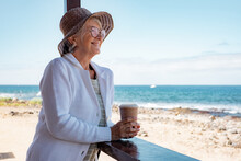 Smiling Senior Woman In Casual Clothing With Hat And Eyeglasses Relaxed At The Beach Looking Away Holding Coffee Cup Enjoying Free Time And Vacation - Horizon Over Sea