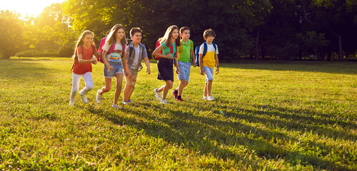 Wall Mural - Happy children walking on green park lawn, enjoying free time and good sunny summer weather. Group of cheerful classmates with rucksacks playing, having fun and exploring nature on school field trip