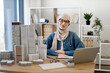 © sofiko14 - Young muslim woman wearing hijab and spectacles sitting at office desk in real estate agency. Efficient realtor using wireless technologies while arranging purchase of residential properties indoors.