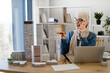 © sofiko14 - Happy muslim woman wearing hijab and spectacles having phone conversation while scanning building models on office desk. Experienced concept architect transferring information to team members.