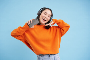 Wall Mural - Beautiful happy korean woman in headphones, listening music on mobile phone in earphones, wearing orange sweatshirt, blue background