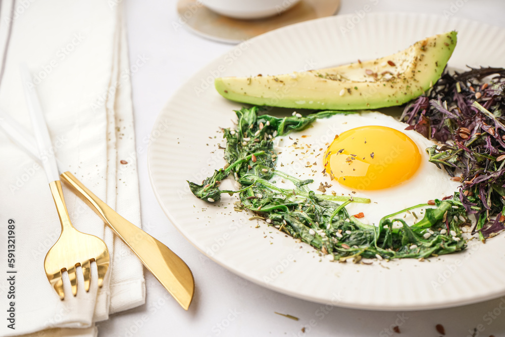 Plate with tasty fried egg, avocado and greens on light background