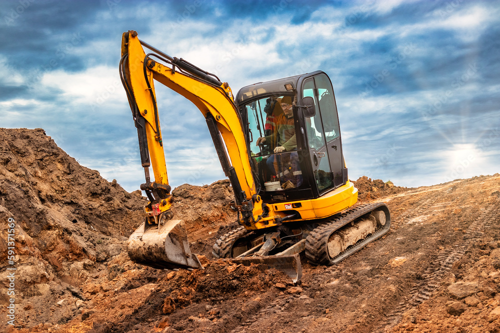 Mini excavator at the construction site on the edge of a pit against a cloudy blue sky. Compact construction equipment for earthworks. An indispensable assistant for earthworks.