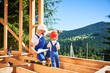 Father with toddler son building wooden frame house. Male builder showing his son the construction plan, wearing helmets and blue overalls on sunny day. Carpentry and family concept.