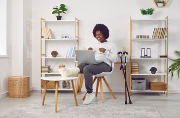 Wall Mural - Smiling African American woman with fractured leg talking on video call. Cheerful beautiful girl with plastered leg sitting on chair using laptop computer at home in living room